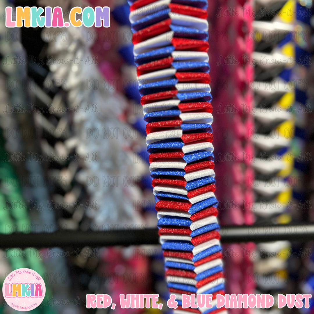 A close-up of a striped fabric trim or ribbon with alternating red, white, and blue glittery textured stripes, hanging vertically against a blurred background of other colorful fabrics. Text overlays for 'LMKIA.